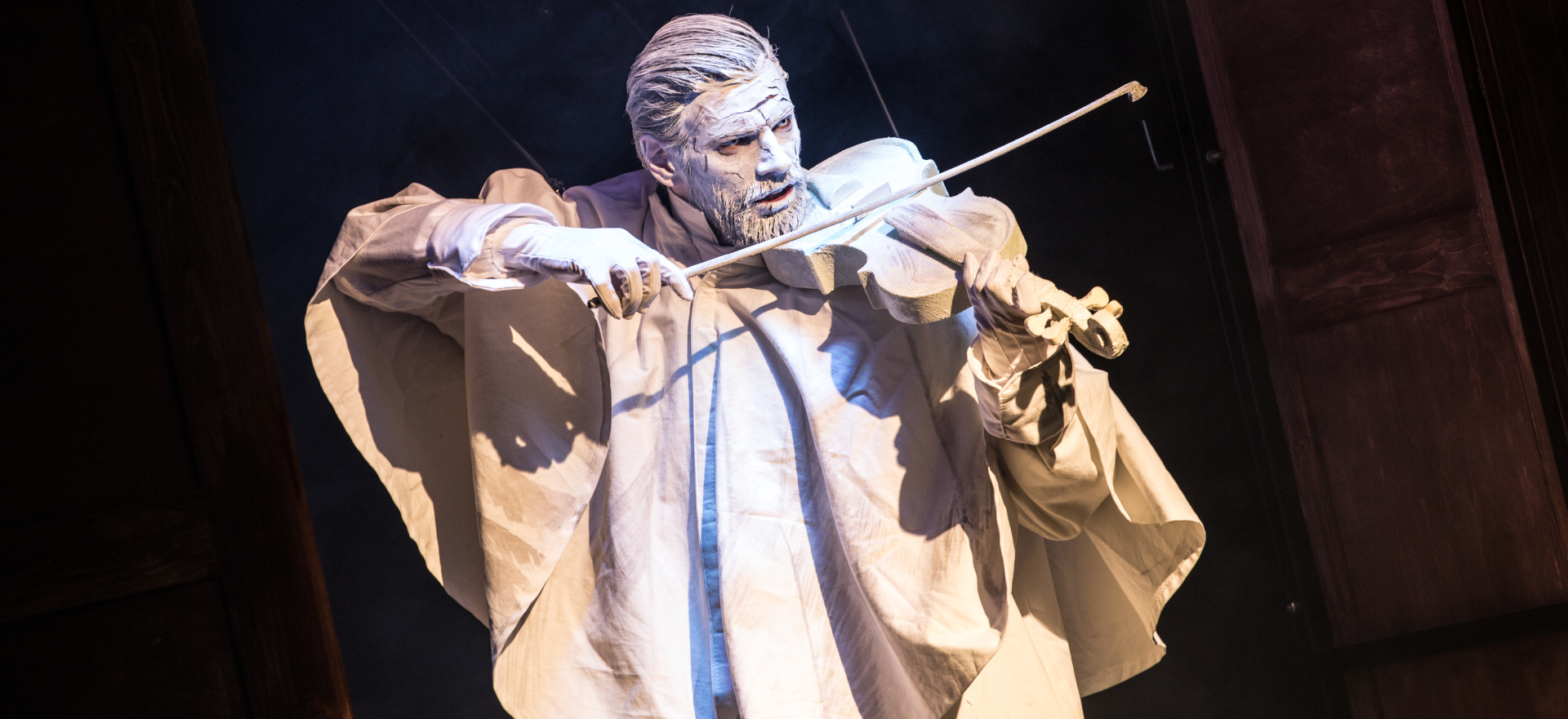mężczyzna w białym płaszczu z pomalowaną na biało twarzą i włosami gra na białych skrzypcach