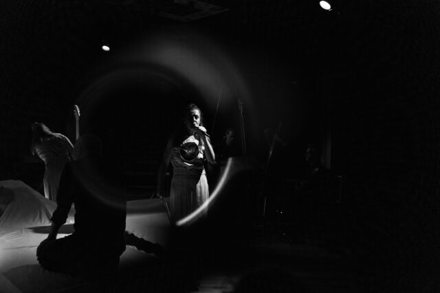 W ciemności na środku widać kobietę w jasnym stroju śpiewająca do mikrofonu. W głębi po lewej niewyraźna postać.